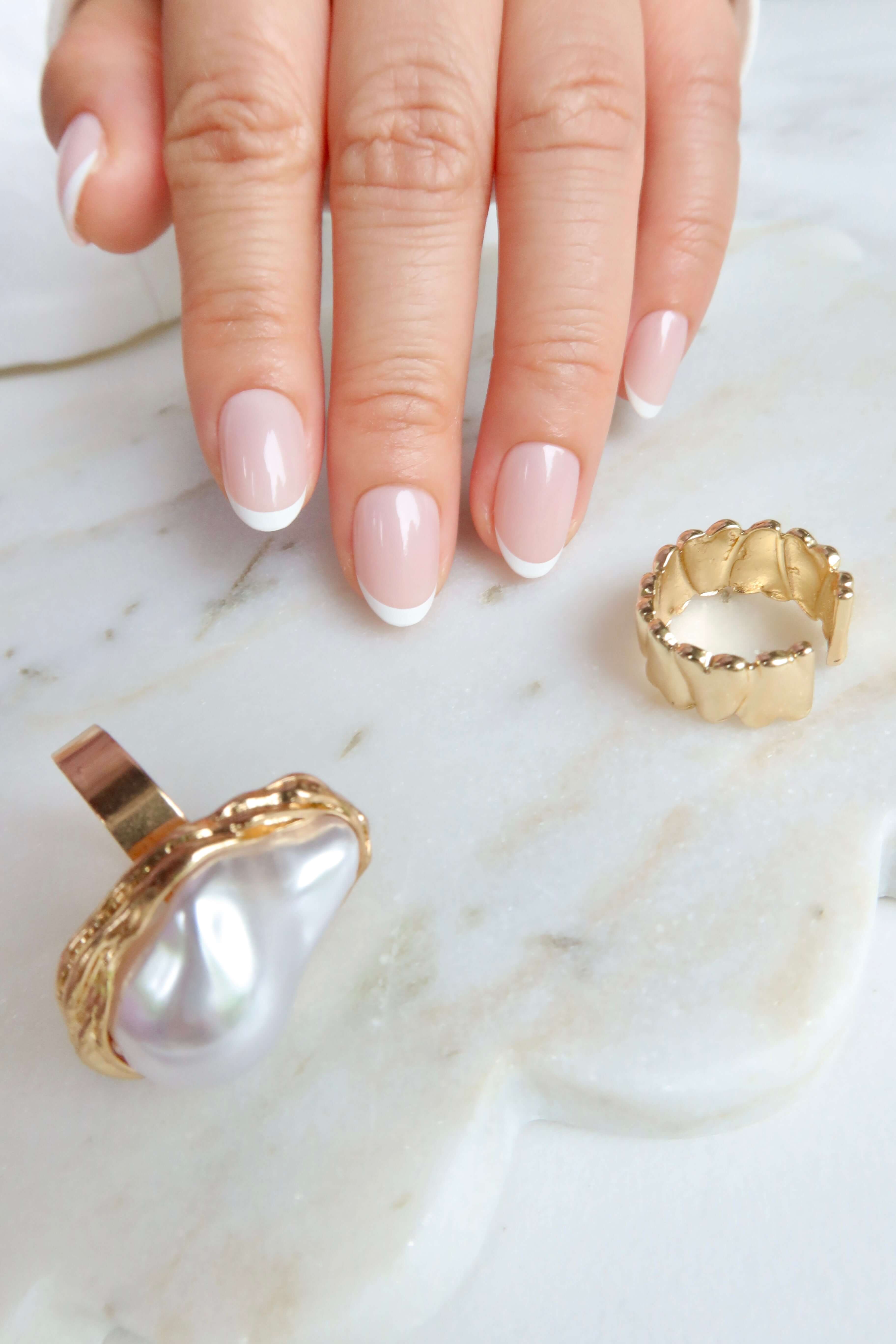 minimal white french tip. short nails. salon quality premier press onsminimal french tip short nails A hand with manicured nails rests on a marble surface, showcasing a large pearl ring and a gold scalloped ring.