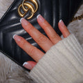 short square french tip nails. press on nails new zealand.Black quilted clutch with a gold logo, resting on a fluffy surface, with a hand displaying French manicure.