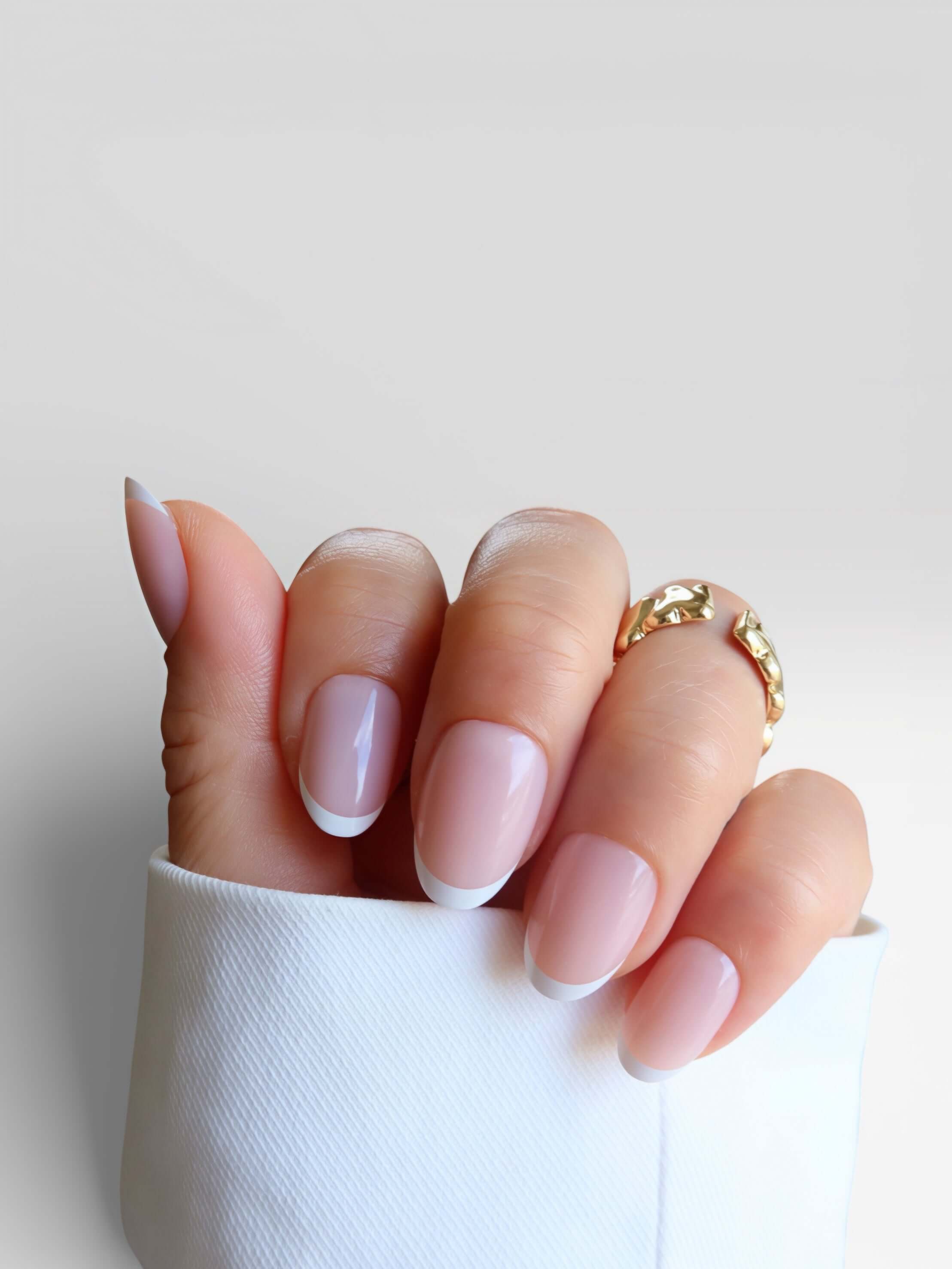minimal white french tip. short nails. salon quality premier press onsLa Parisienne manicure featuring light pink nails with white tips, hand adorned with a gold ring against a white background.